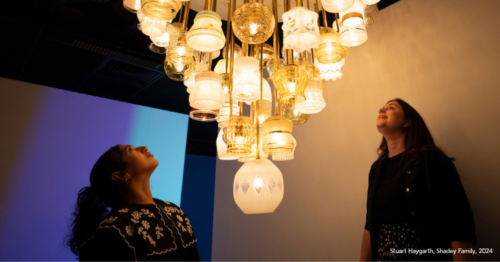 Two woman stood looking upwards at a light artwork made of different glass lamps inside The Light Durham.
